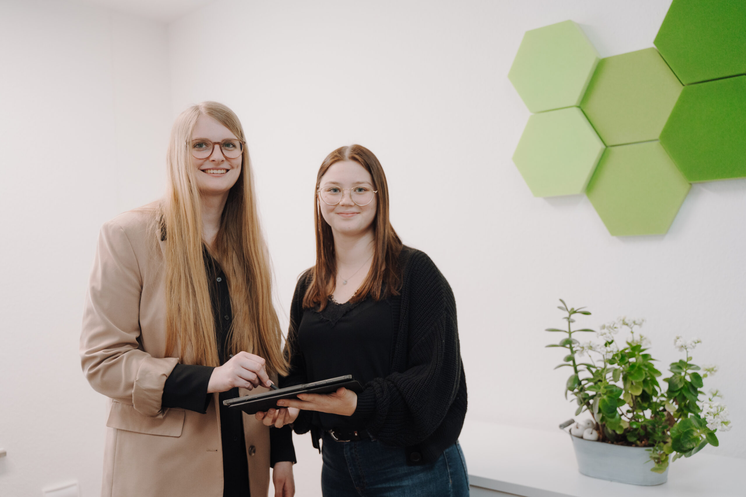 zwei frauen halten gemeinsam ein tablet und stehen vor einer wand mit gruenen sechsecken